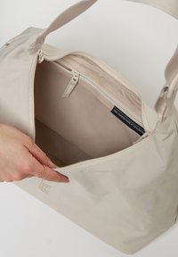 Beige Tasche mit einer glatten, glänzenden Oberfläche aus recycelten Materialien. Das Innere enthält eine Reißverschlusstasche und ein Etikett, das die Materialzusammensetzung angibt.
