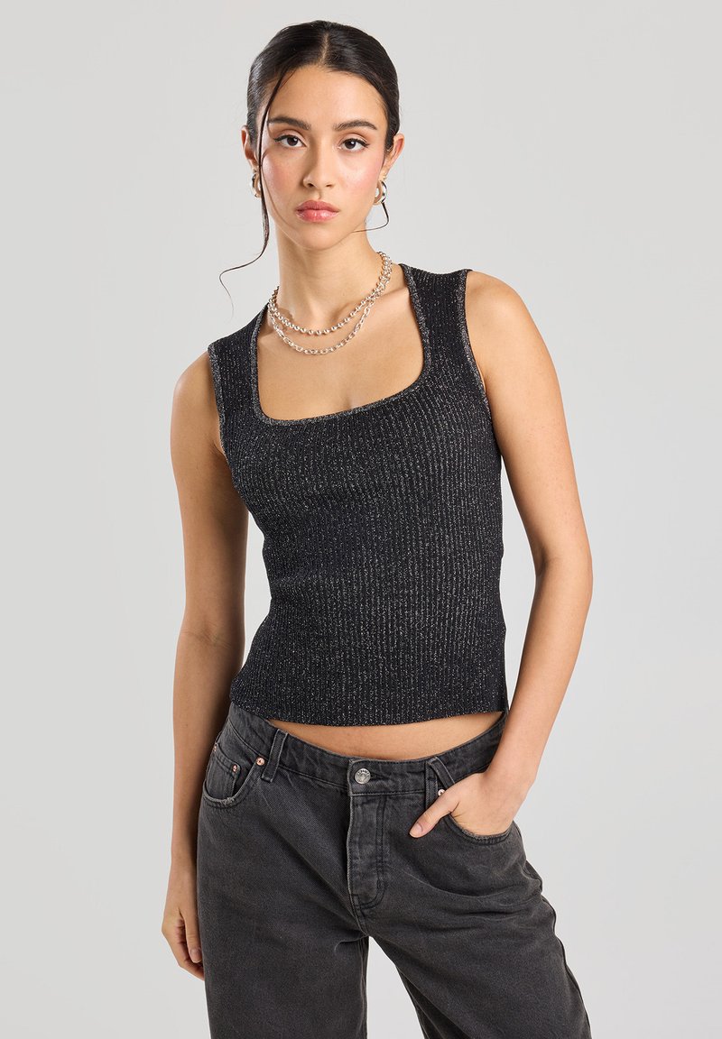 Haut sans manches noir à côtes avec un décolleté carré, ornée de fils métalliques. Porté avec un pantalon décontracté gris foncé.