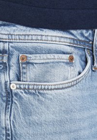 Light blue denim jeans feature a rear pocket with copper rivets, contrasting stitching, and a smooth texture, with a dark waistband visible above.