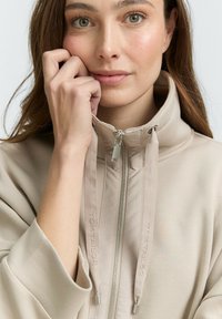 Žena s svijetlosmeškom kosom koja nosi bež zip-up jaknu s visokim ovratnikom i vezicama, dodiruje svoje lice desnom rukom.