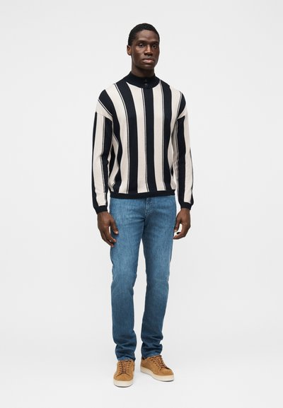 Oprijet pulover z obrnjenimi mornarsko modrimi in kremnimi črtami, visokim ovratnikom in detajlom z gumbi. Kombiniran z svetlo modrimi kavbojkami in čevlji v bež barvi.