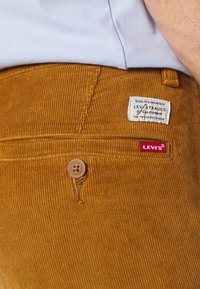 Gros plan sur la poche d'un pantalon Levi's en velours côtelé moutarde avec un bouton et deux étiquettes de marque en tissu, porté par une personne portant une chemise bleu pâle.