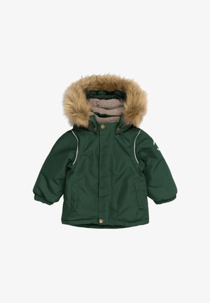 Un niño sentado sobre un tronco, vestido con una chaqueta acolchada verde oscuro con capucha forrada de piel y pantalones de esquí azules con estampado, con las manos apoyadas sobre el tronco.