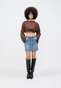 Bluză scurtă maro cu design în relief, combinată cu o fustă mini din denim cu nasturi în față și cizme negre până la genunchi cu toc.