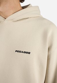 Beige színű kapucnis pulóver, texturált anyagból, a mellrészen hangsúlyos fekete "PEGADOR" logóval. Egyszerű dizájn, minta nélkül.
