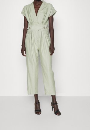 Personne portant une combinaison ceinturée vert clair à manches courtes, associée à des sandales noires à talons hauts, debout devant un fond uni.