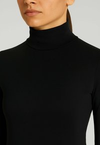 Donna con un maglione aderente nero a collo alto e maniche lunghe, mostrata dalla zona del collo fino all'altezza del busto su uno sfondo semplice.