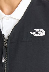 Chaqueta negra con cremallera y textura acanalada, con un logo bordado en blanco en el pecho y una camiseta polo blanca debajo.