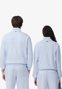 Felpa azzurra chiara con colletto alto e polsini a costine. Presenta un logo verde sulla schiena. Tessuto morbido e vestibilità comoda.