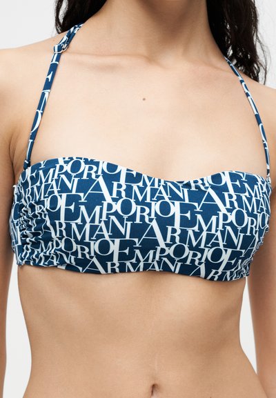 Μπλε μπαντό μαγιό με λευκό κείμενο "EMPORIO ARMANI" σε όλο το ύφασμα, φορεμένο από ένα άτομο με μακριά σκούρα μαλλιά.