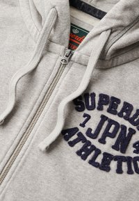 Felpa grigia con zip in pile, con scritta ricamata in blu navy "SUPERDRY JPN ATHLETIC" sul davanti e cappuccio con coulisse.