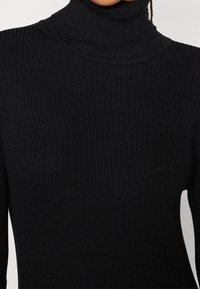 Maglione a collo alto di colore nero, realizzato in tessuto a costine. Presenta un design aderente e un collo alto. Texture liscia con un colore uniforme.