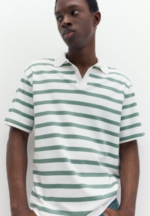 Jeune homme portant un polo blanc à rayures horizontales vertes, debout devant un fond blanc uni.