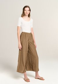 White short-sleeve bodysuit paired with loose, crinkled brown wide-leg pants. Shoes are light brown slide sandals with a braided strap.
