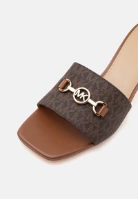 Sandale à enfiler marron avec bout carré, large bride à motif et détail du logo MK circulaire doré sur le dessus.
