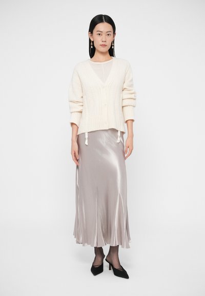 Krémový svetr s kabelovým vzorem a výstřihem do V s třásněmi, nošený přes béžovou midi sukni z hedvábí, doplněný černými lodičkami na jehlovém podpatku.