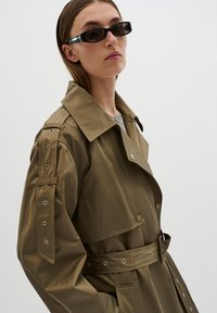 Cappotto trench verde oliva con ampio colletto, vita regolabile con cintura e occhielli alle maniche. Modella che indossa occhiali da sole rettangolari.