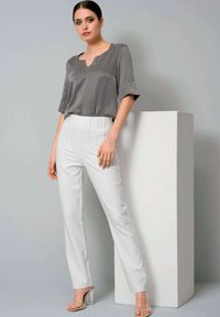 Gray, short-sleeve blouse with a V-neck; paired with white, tailored pants featuring vertical pleats. Model stands next to a white pedestal.
