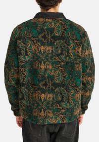 Pluizige jas met groen en bruin patroon en een zwarte kraag. Heeft een zachte textuur en een losse pasvorm, met overal bloemmotieven.