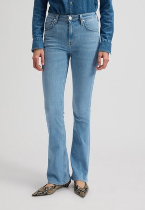 Personne portant un jean évasé bleu clair, une chemise en denim foncé boutonnée, et des ballerines pointues à motif peau de serpent.