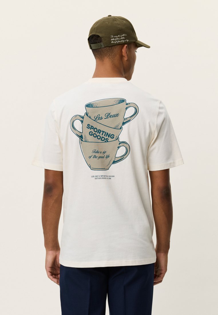 Λευκό βαμβακερό μπλουζάκι με γραφικό στοιχείο στοιβαγμένων φλιτζανιών καφέ, τιρκουάζ κείμενο που αναγράφει "Les Deux" και "SPORTING GOODS", με casual εφαρμογή.