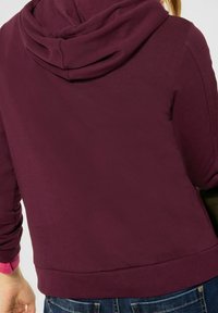 Bordowa bluza z kapturem o miękkiej fakturze, z kieszenią kangurką, ściągaczami na mankietach i u dołu. Widok z tyłu pokazuje detale kaptura.