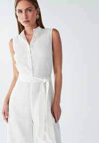 Jumpsuit blanco sin mangas con cuello, cierre de botones y cinturón de tela. Textura suave, corte relajado y diseño de pierna ancha.