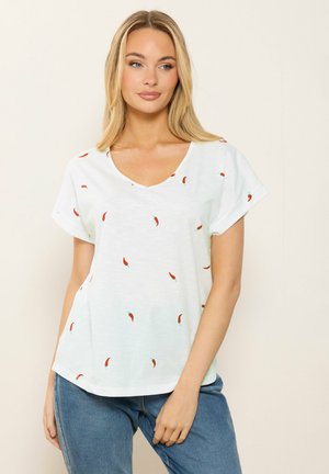 Femme aux longs cheveux blonds portant un T-shirt blanc à col en V avec des imprimés de piments rouges et un jean bleu, debout devant un fond clair uni.