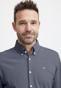 Camicia grigia da uomo con colletto e bottoni bianchi. Presenta un tessuto strutturato e un piccolo logo ricamato in rosso, bianco e blu.