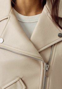 Giacca beige in ecopelle con cerniera argentata, bottoni a pressione e colletto a lancia; tasche visibili. Tessuto strutturato con finitura morbida.