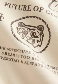 Kreemivärvi kangas, millel on tiigri illustratsioon ümmarguse köite kujunduses, mille kõrvale on lisatud dekoratiivne tekst tumedat pruunis.