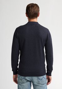 Man met zijn rug naar de camera, gekleed in een donkerblauwe trui en lichte spijkerbroek, tegen een eenvoudige lichte achtergrond.