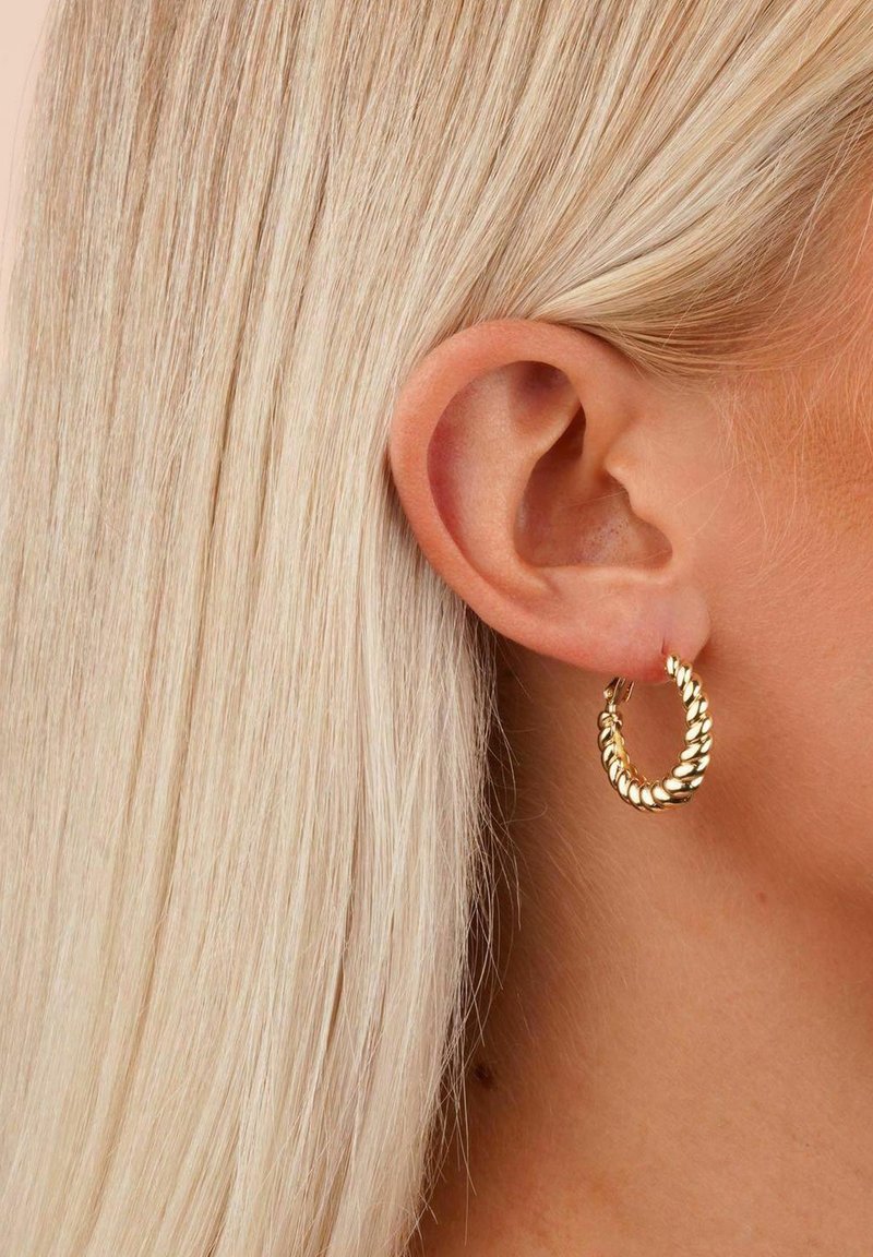 Goldene Creolen mit einem gedrehten Design, das eine glänzende Oberfläche und einen sicheren Verschluss aufweist, getragen am Ohr einer Person mit glattem, blonden Haar.