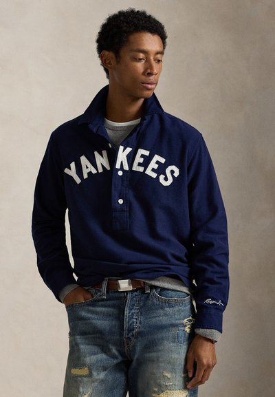 Polo de manga longa da marinha com o logótipo "YANKEES", placket de dois botões, punhos cinza e jeans de ganga desbotados com desgaste e um cinto castanho.