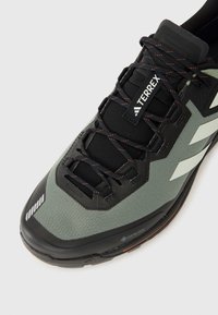 Črne in zelene pohodniške čevlje z mrežastim zgornjim delom, teksturirano blago, ojačano konico in črnimi vezalkami. Imajo vidno oznako GORE-TEX.