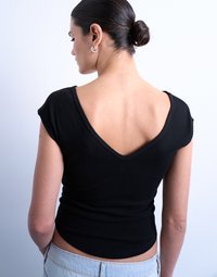 Γυναίκα με τα μαλλιά της σε κότσο, που φοράει κρίκους στα αυτιά, μαύρο τοπ με V-πλάτη και ανοιχτόχρωμα τζιν, που φαίνεται από πίσω μπροστά σε ένα απλό ανοιχτόχρωμο φόντο.