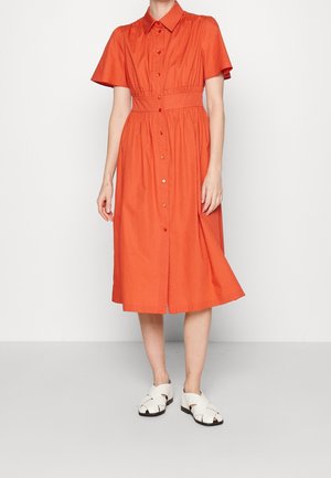 Kvinde iført en orange knælængde kjole med knapper, korte ærmer og tilspidset talje, kombineret med hvide åbne sandaler.