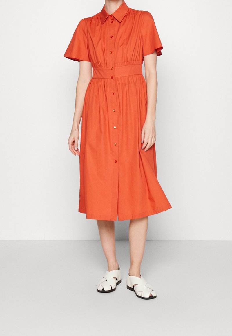 Femme portant une robe boutonnée orange jusqu'aux genoux avec des manches courtes et une taille cintrée, associée à des sandales blanches à bouts ouverts.