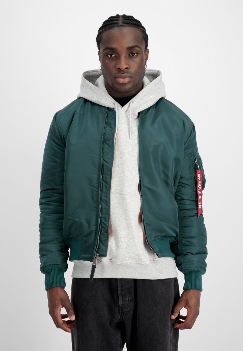 Giacca bomber verde scuro realizzata in materiale liscio con polsini a costine, abbinata a un hoodie grigio e jeans neri. Presenta un'etichetta rossa sulla manica.