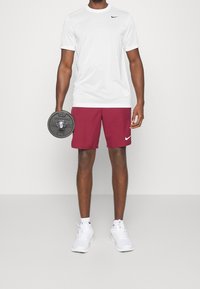 Camiseta blanca de manga corta de Nike combinada con pantalones cortos color burdeos. La persona sostiene un peso negro, viste zapatillas blancas y calcetines tobilleros.
