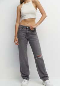 Jean gris clair taille haute avec une coupe ample, présentant une grande déchirure au niveau du genou. Porté avec un débardeur court côtelé et des baskets blanches.