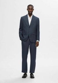 Abito blu scuro realizzato in tessuto testurizzato, vestibilità su misura con giacca a un bottone, camicia bianca sotto e scarpe nere.