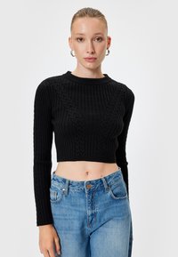Zwarte cropped trui met ribbelstructuur en kabelbreisel; lange mouwen en ronde halslijn. Gepaard met lichtblauwe jeans.