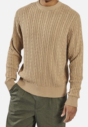 Mann trägt einen beige gestrickten Zopfpullover mit gerippten Bündchen und Saum, kombiniert mit olivgrünen Hosen, die Hände in den Taschen.
