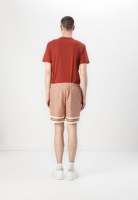 Rød bomulls t-skjorte kombinert med mønstrete oransje shorts med hvite og røde striper. Modell iført hvite sneakers og synlige ankler.