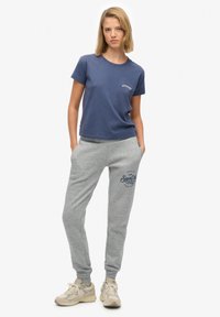 Camiseta de algodón azul marino de mangas cortas con logo blanco, combinada con pantalones de chándal grises con cordón y diseño impreso. Zapatillas beige elegantes.