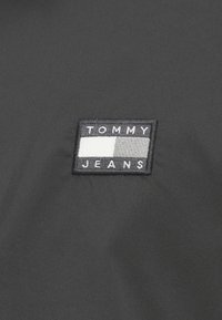 Musta kangas, jossa on suorakulmainen brodeerattu päällyste, jossa lukee "TOMMY JEANS" valkoisella mustaa ja harmaata taustaa vasten.