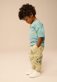 Fiatal gyermek göndör hajjal, kék pólót, bézs munkanadrágot traktor folttal és fehér sportcipőt visel, lefelé néz, miközben a zsebébe dugja a kezét.