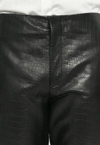 Pantaloni in ecopelle di coccodrillo nera indossati sopra un tessuto bianco, che mostrano una texture dettagliata e cuciture centrali.