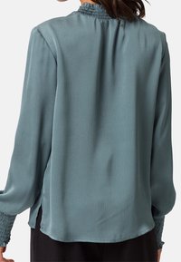 Blouse à manches longues couleur sarcelle en tissu texturé, avec un col et des poignets smockés, et des fentes latérales pour le style et le confort.